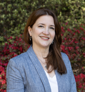 Headshot of Dr. Laila Ferreira wearing a blue blazer in front of red and green foliage